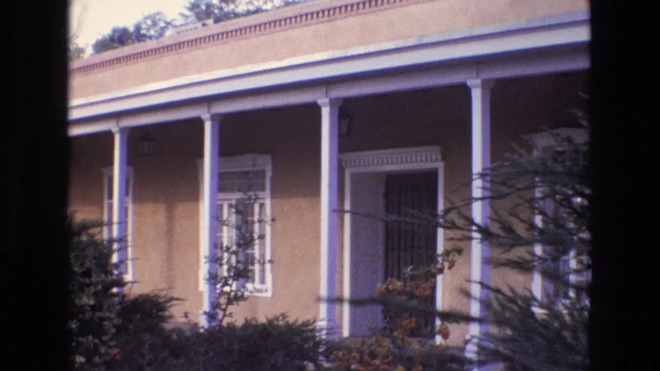 New Mexico 1973 home-movie frame showing an adobe-style building with white porch columns, barred windows, foreground foliage, and part of the street.