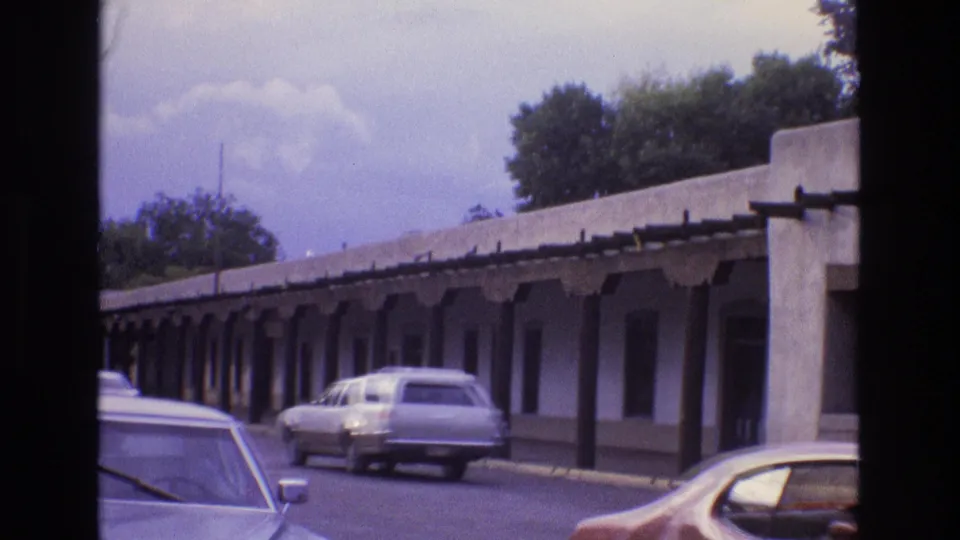 New Mexico 1973 street view with a long covered adobe-style walkway, dark support posts, a white station wagon, and other cars near the curb.