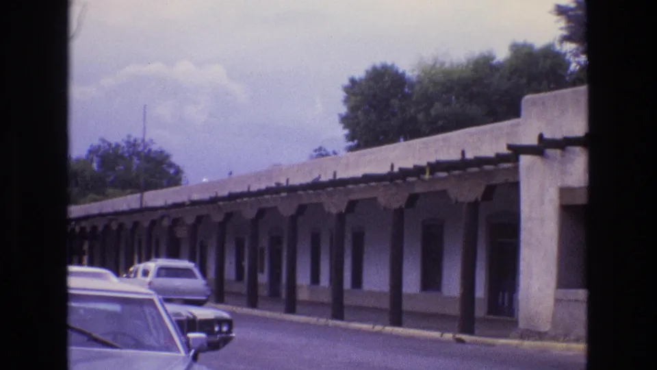 New Mexico 1973 home-movie frame showing a long adobe-style portico, repeated dark columns, street trees, and cars positioned along the roadway.