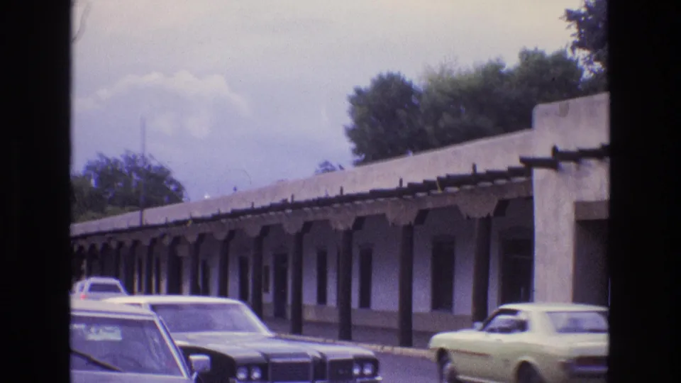 New Mexico 1973 street scene with an adobe-style covered walkway, parked and passing classic cars, trees, and a pale sky in soft 8mm color.