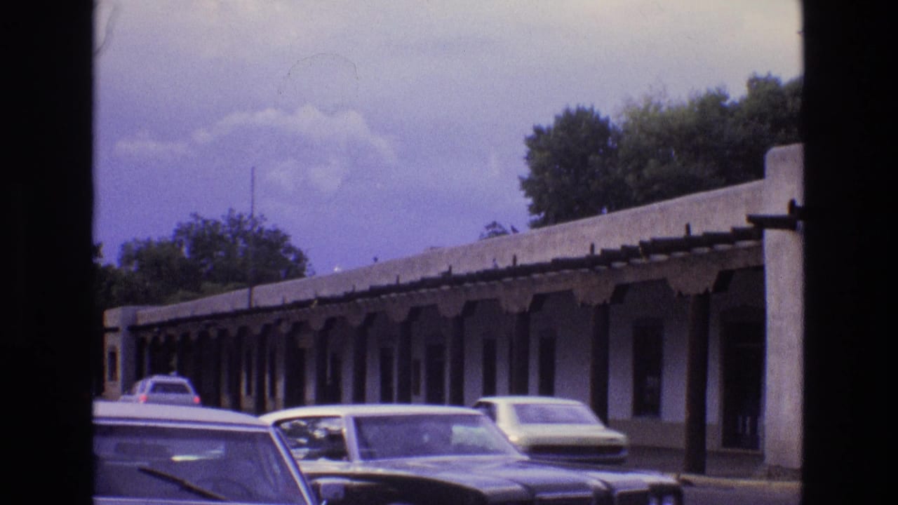 Cars pass a long adobe-style building with a shaded covered walkway.
