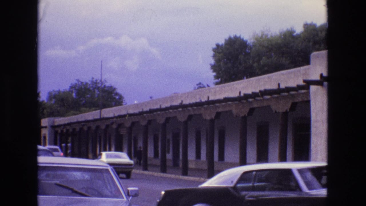 1973 New Mexico street view with classic cars passing a long adobe-style frontage and shaded walkway.
