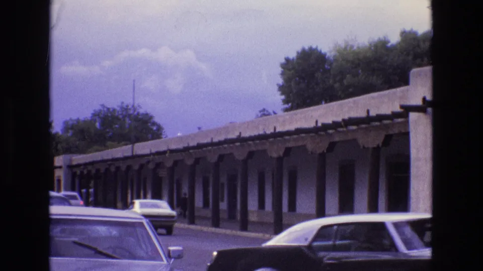 1973 New Mexico street view with classic cars passing a long adobe-style frontage and shaded walkway.