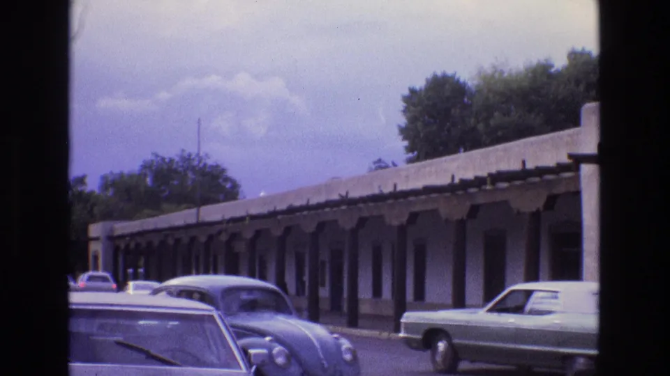 1973 New Mexico home-movie frame with multiple cars along a street beside a long adobe-style building and shaded portal.