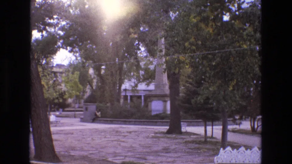 1973 New Mexico travel footage shifts to a quiet park-like area with mature trees, a walkway, open ground, and a small monument or sign.