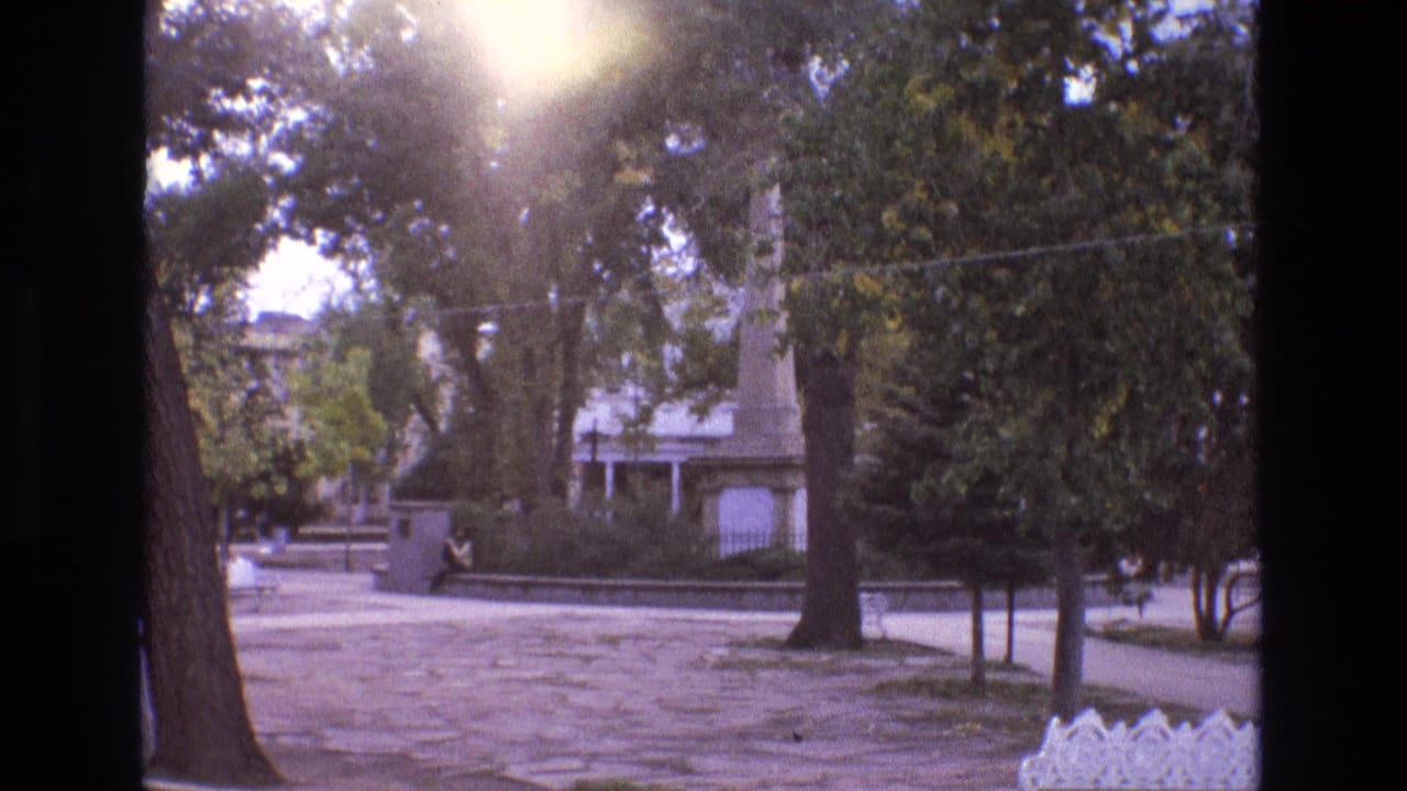 1973 New Mexico 8mm frame showing a sunlit park or plaza with mature trees, paved foreground, a low wall, a distant seated person, and a vertical monument-like structure.