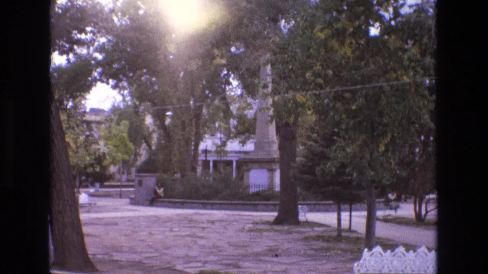 1973 New Mexico 8mm frame showing a sunlit park or plaza with mature trees, paved foreground, a low wall, a distant seated person, and a vertical monument-like structure.