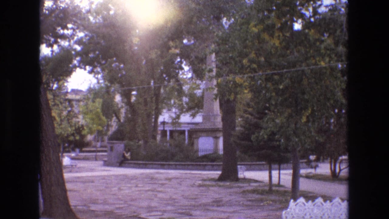 1973 New Mexico home-movie frame of a tree-lined park area with strong sun flare, stone paving, a low wall, a distant seated figure, and a monument-like vertical feature.