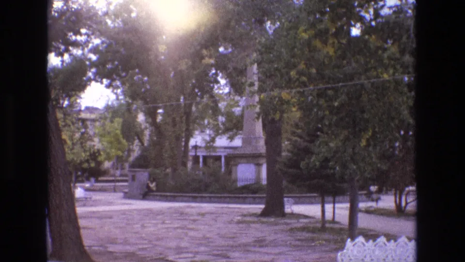 1973 New Mexico home-movie frame of a tree-lined park area with strong sun flare, stone paving, a low wall, a distant seated figure, and a monument-like vertical feature.