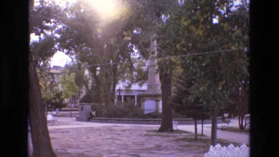 1973 New Mexico 8mm frame showing the same sunlit park view with soft focus, tree trunks, paved ground, a low wall, a distant seated person, and a monument partly framed by foliage.