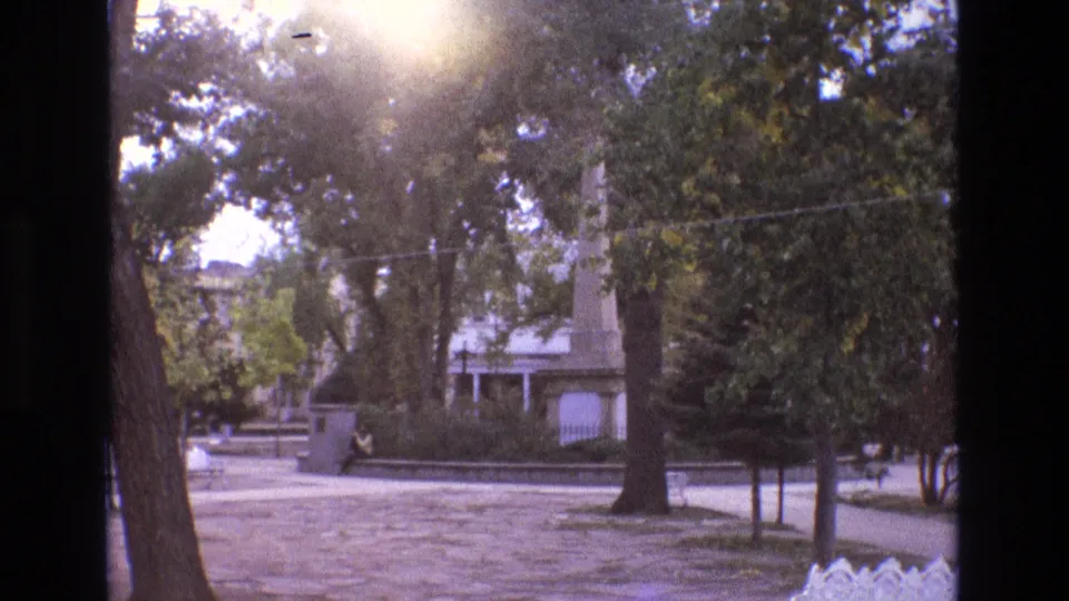 1973 New Mexico 8mm frame of a bright landscaped park or plaza, with mature trees, strong sun flare, a vertical monument, low wall, paved foreground, and a distant seated person.