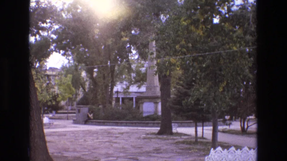 1973 New Mexico home-movie frame showing a quiet park with mature trees, a paved walkway, white metal benches, distant structures, and a small distant person near the left side.