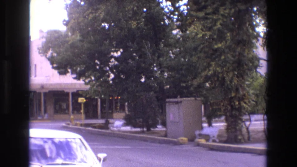 1973 New Mexico 8mm frame captured from a moving vehicle, with a white car in the foreground and a tree-lined urban street, benches, utility box, and low-rise building beyond.