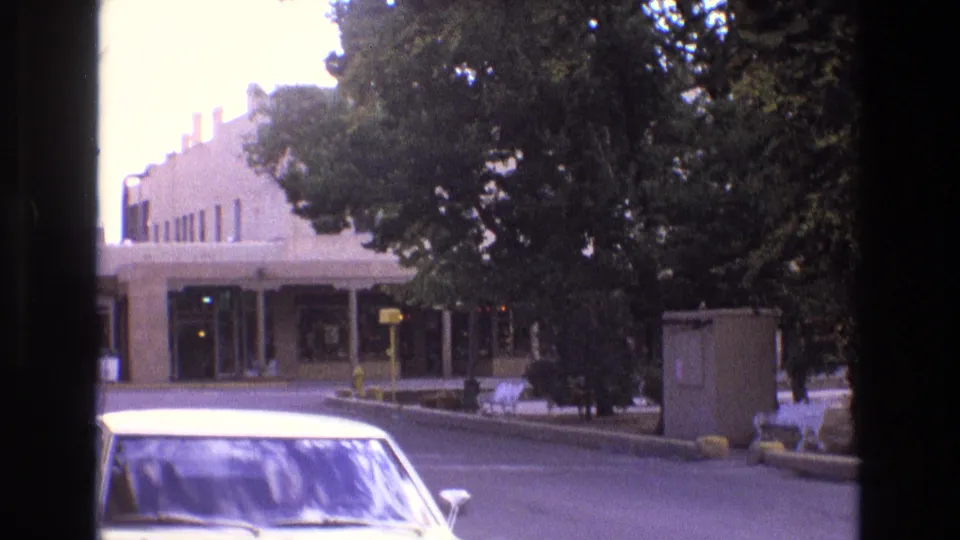 1973 New Mexico 8mm frame showing the front portion of a white car beside a quiet street with mature trees, white benches, a beige utility box, and a light-colored building with large windows.