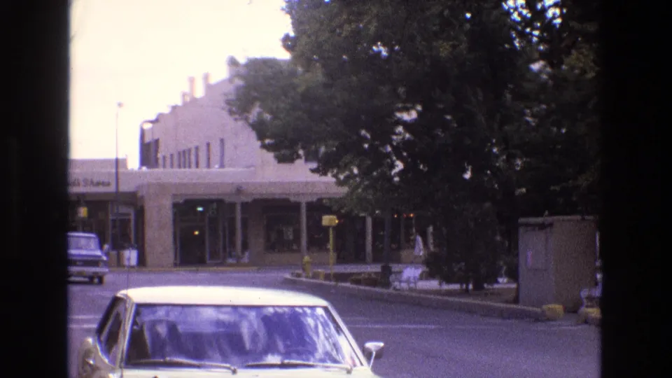 1973 New Mexico street view with a light-colored sedan in the foreground, a low-rise commercial building across the road, trees, signs, and a parked vehicle beyond.