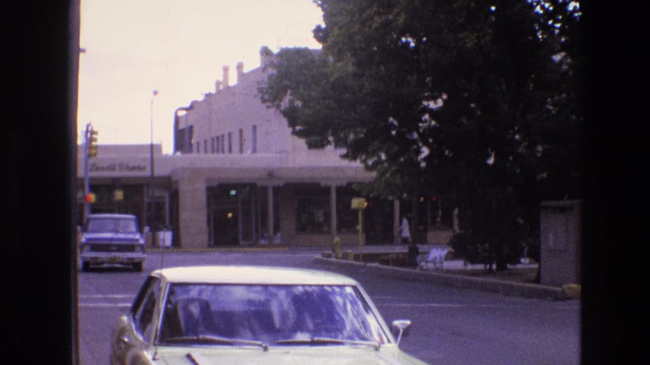 A light-colored 1970s-era sedan occupies the foreground while a mid-century commercial frontage, trees, street signs, and a boxy parked vehicle sit across the street.