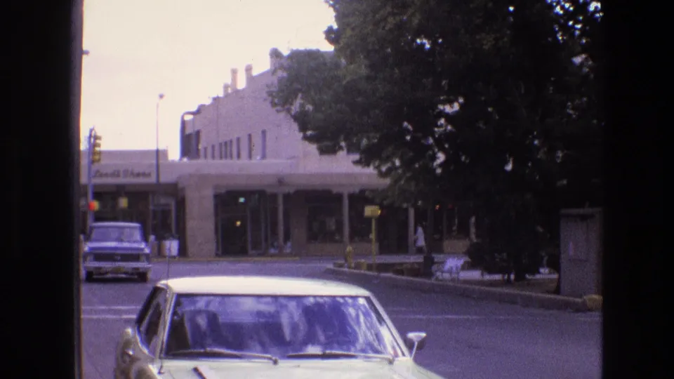 A light-colored 1970s-era sedan occupies the foreground while a mid-century commercial frontage, trees, street signs, and a boxy parked vehicle sit across the street.