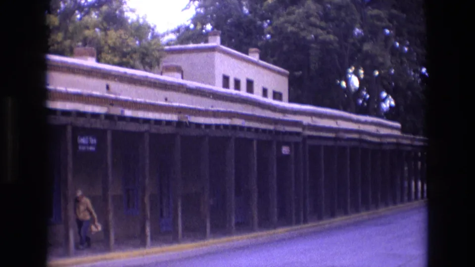 1973 New Mexico home-movie frame showing a long covered walkway or storefront arcade, shaded columns, street pavement, trees, and a pedestrian walking near the left side.