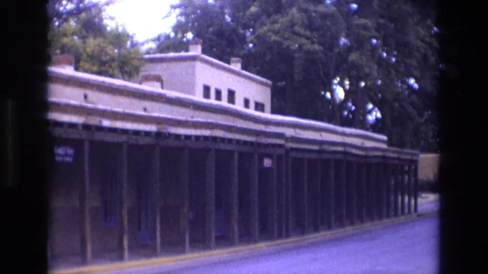A long shaded storefront walkway in New Mexico appears beside an empty street, with columns, upper building windows, trees, and faded 8mm color visible in the frame.