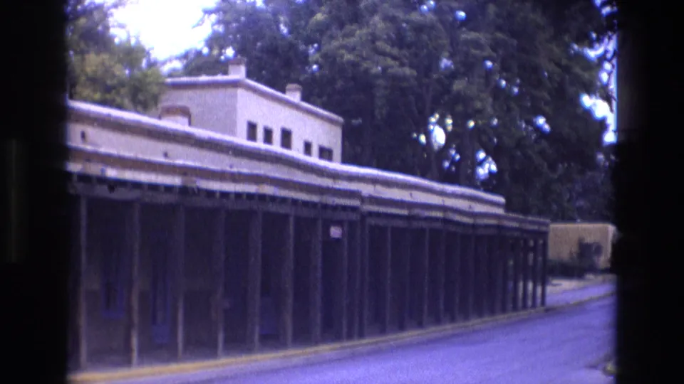 1973 New Mexico home-movie frame showing a long low adobe-style building with a covered arcade beside a paved street, partly framed by a dark vehicle interior.