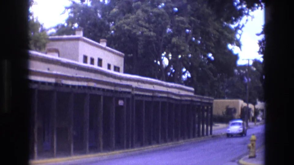 1973 New Mexico street view with a white Volkswagen Beetle approaching past a long adobe-style building, trees, and a yellow fire hydrant.