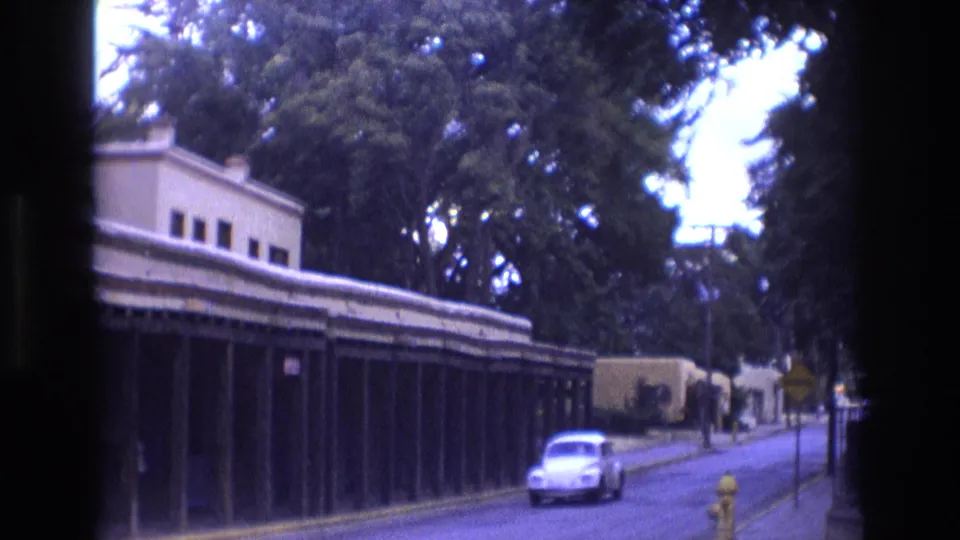 1973 New Mexico home-movie frame of a white Volkswagen Beetle passing a long adobe-style arcade building on a quiet paved street.