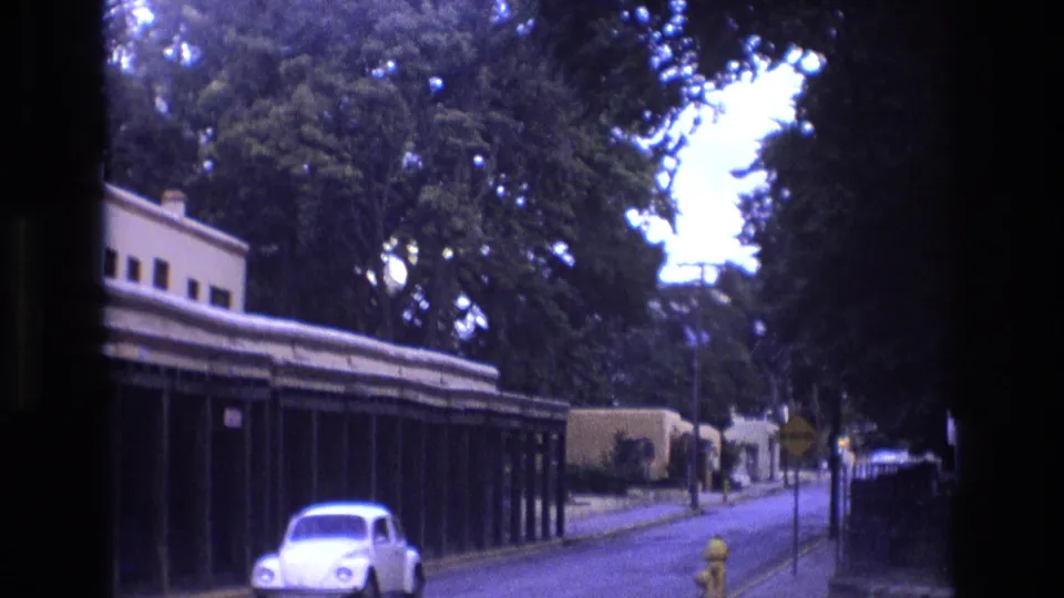 1973 New Mexico street frame with a white Volkswagen Beetle near the foreground, a yellow fire hydrant, and a long adobe-style building under mature trees.
