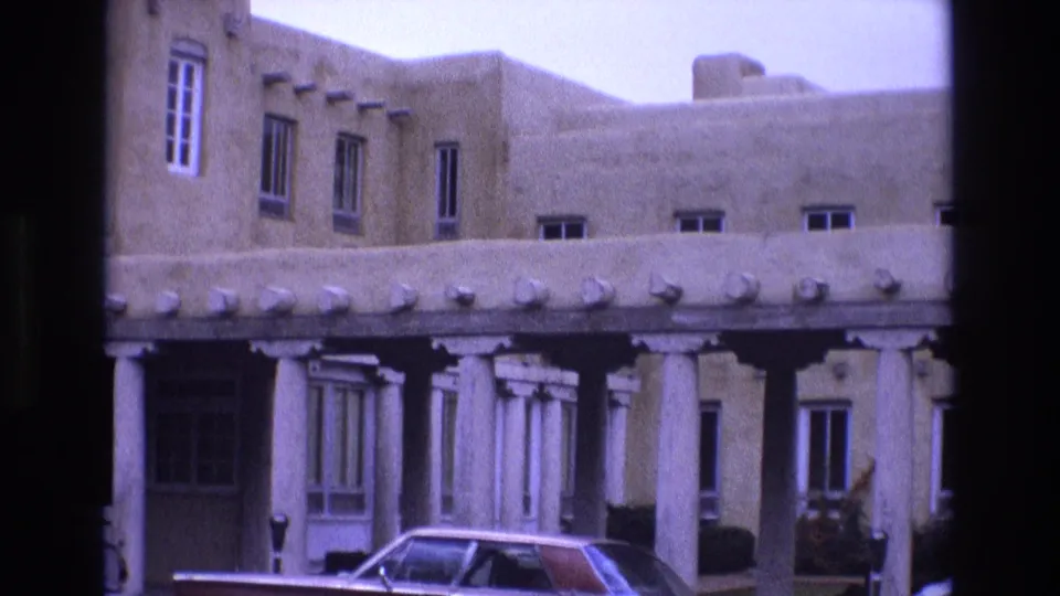 1973 New Mexico home-movie frame showing an adobe-style building facade with white columns, a covered breezeway, and a parked car at street level.