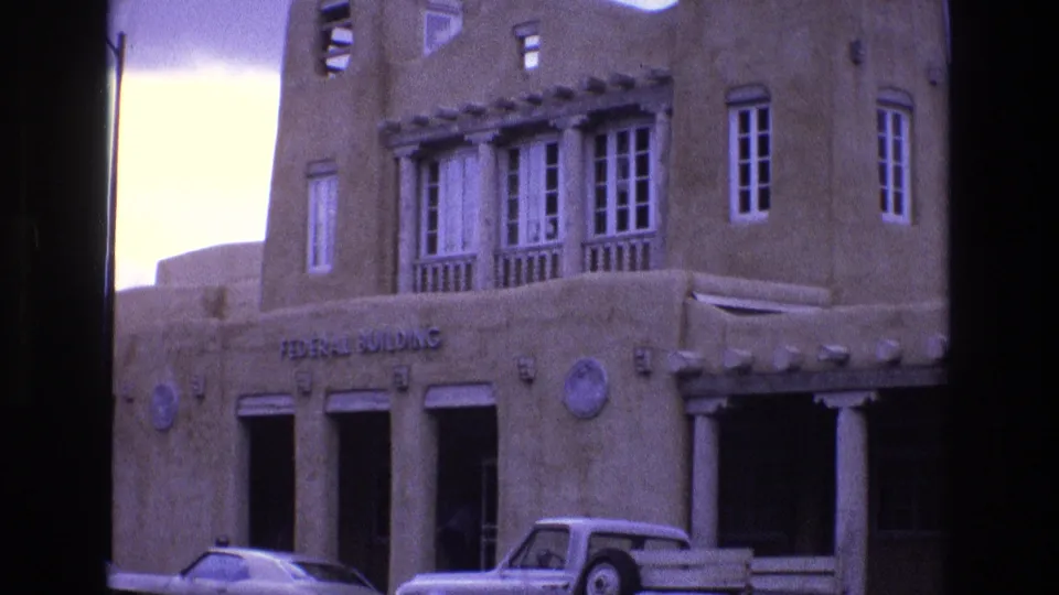 1973 New Mexico home-movie frame showing an adobe-style Federal Building facade with white-framed windows, columns, and parked period cars.