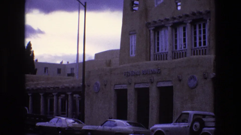 Home-movie frame from 1973 New Mexico showing part of the Federal Building facade, parked cars, a streetlight, and pronounced dark frame borders.