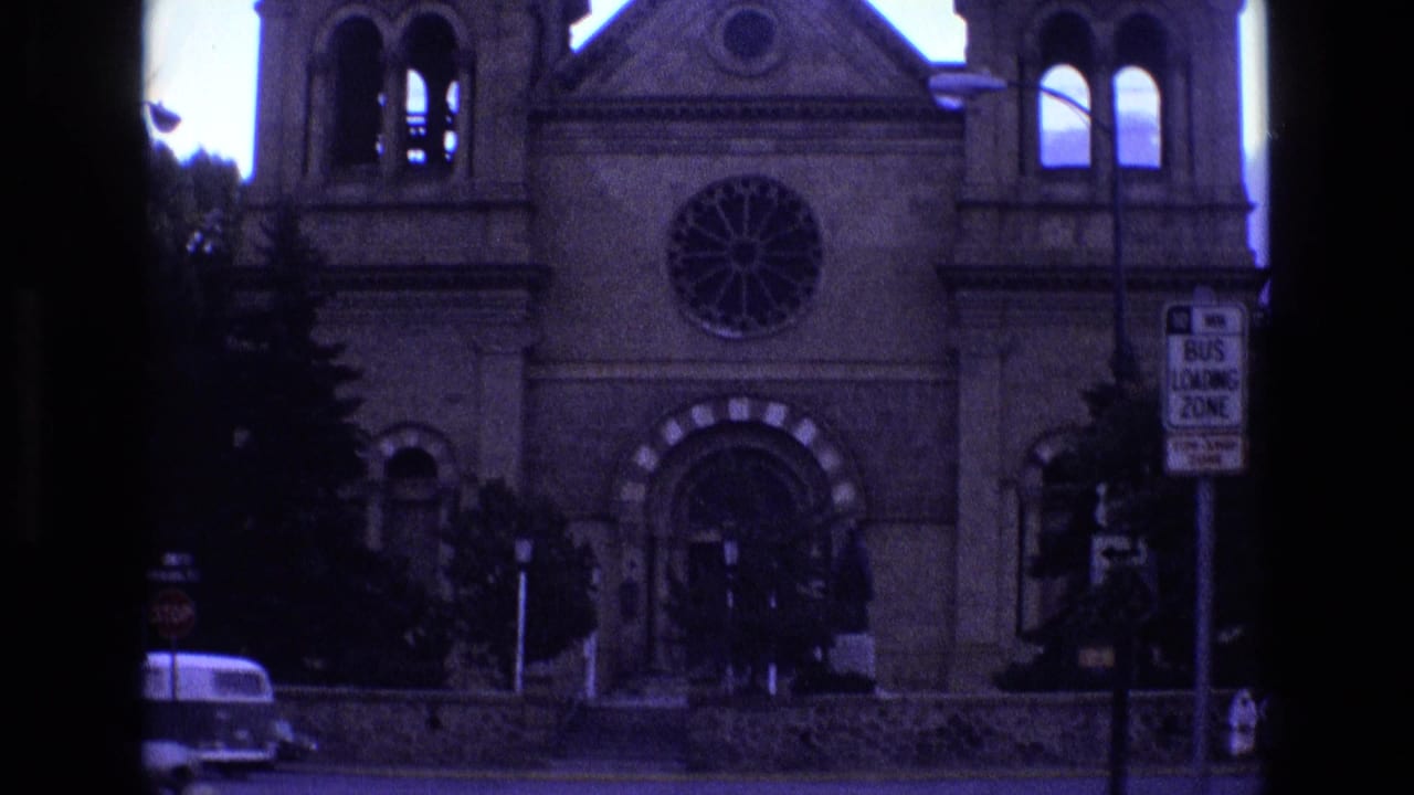 1973 New Mexico 8mm frame showing a stone church or cathedral facade with twin towers, a circular rose window, a parked vehicle, and a bus loading zone sign across the street.