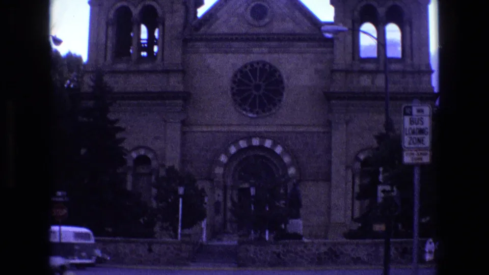 1973 New Mexico 8mm frame showing a stone church or cathedral facade with twin towers, a circular rose window, a parked vehicle, and a bus loading zone sign across the street.