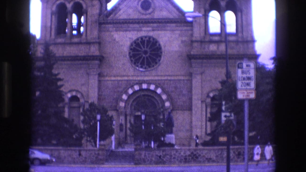 1973 New Mexico 8mm frame of a historic stone church facade with a rose window, arched entry, bus loading zone sign, parked cars, and small pedestrians near the sidewalk.