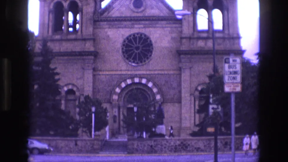 1973 New Mexico 8mm frame showing the church or cathedral front with rose window, striped arched entrance, bus loading zone sign, stone wall, and pedestrians along the sidewalk.