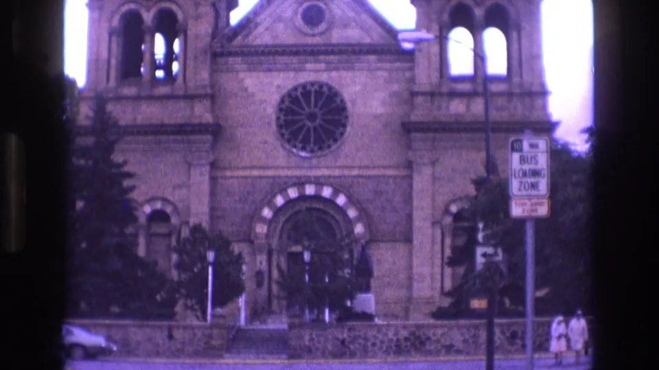 1973 New Mexico 8mm frame holding on the stone church or cathedral facade, with a circular rose window, arched entrance, street signage, a parked car, and pedestrians at the right edge.