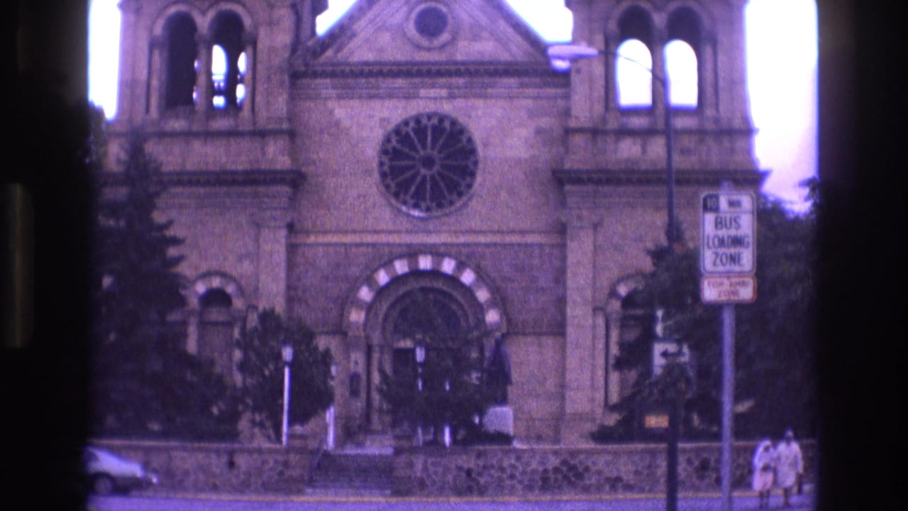 1973 New Mexico 8mm frame showing the Cathedral Basilica of St. Francis of Assisi facade from across the street, with a rose window, arched entry, bus loading zone sign, and pedestrians near the sidewalk.