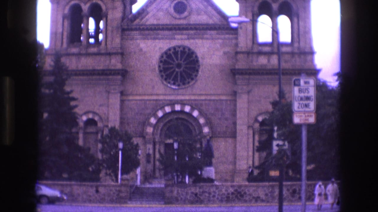 1973 New Mexico 8mm frame of the cathedral exterior, with the round rose window centered above the entrance, twin towers visible, a bus loading zone sign on the right, and pedestrians at the sidewalk edge.