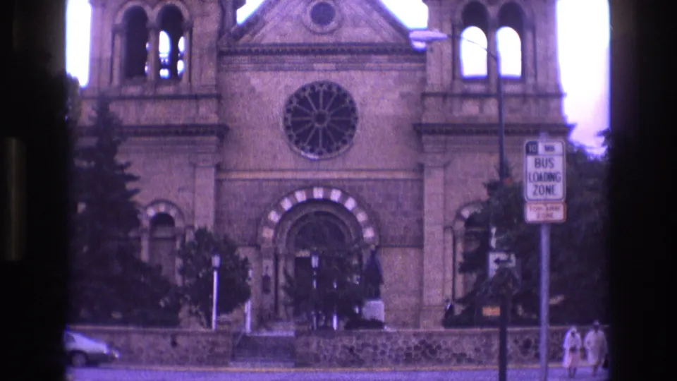 1973 New Mexico 8mm frame of the cathedral exterior, with the round rose window centered above the entrance, twin towers visible, a bus loading zone sign on the right, and pedestrians at the sidewalk edge.
