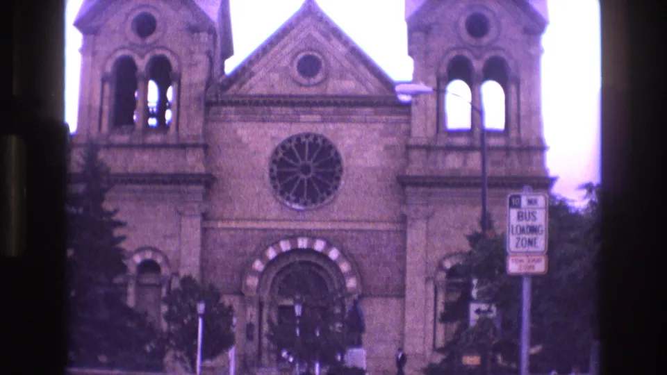 1973 New Mexico home-movie frame showing a tighter view of the Cathedral Basilica of St. Francis of Assisi facade, including the rose window, arched entrance, twin towers, and bus loading zone sign.