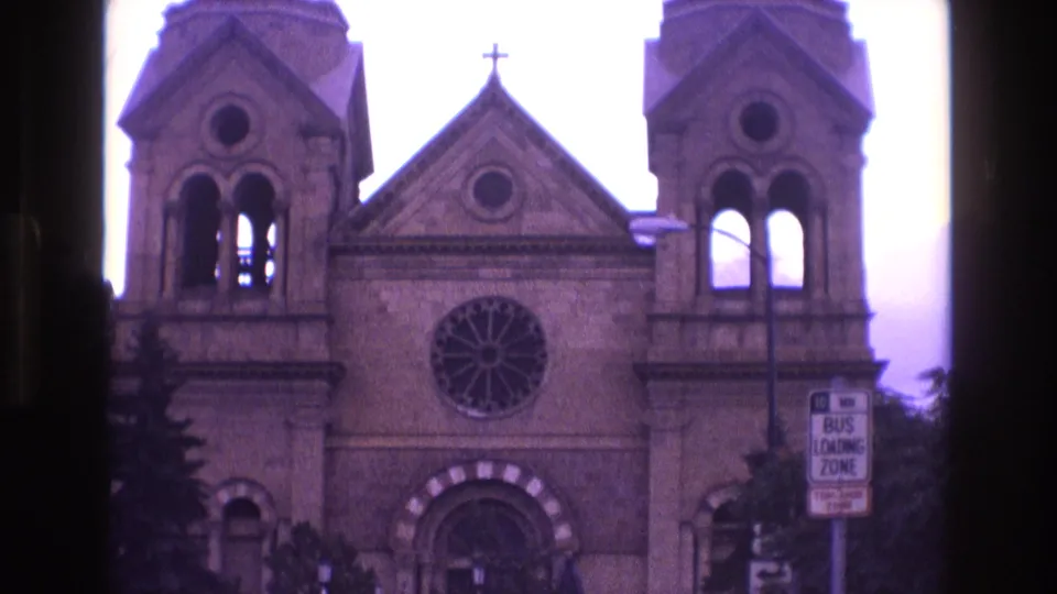 1973 New Mexico 8mm frame of the cathedral facade shifted upward, emphasizing the twin towers, roofline cross, circular rose window, arched entry, and bus loading zone sign near the street.