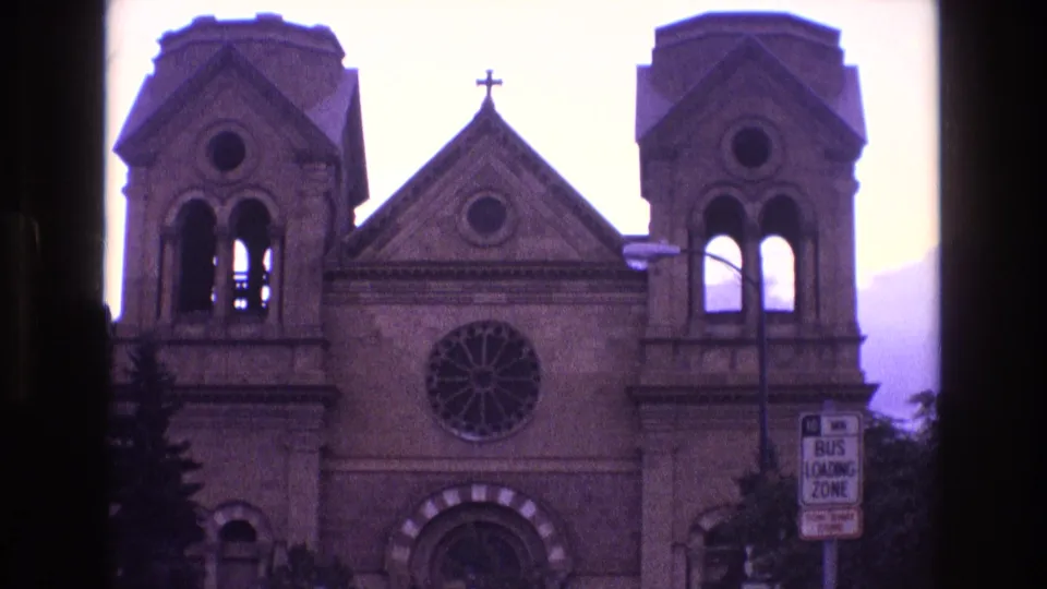 1973 New Mexico 8mm frame showing the front of a church with twin bell towers, a central rose window, a gable cross, trees, and a bus loading zone sign at right.
