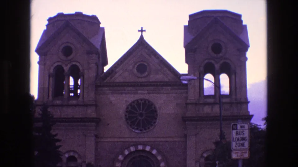 1973 New Mexico 8mm frame with a church facade, twin tower openings, circular rose window, cross on the gable, silhouetted trees, and a bus loading zone sign.