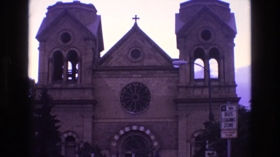 1973 New Mexico 8mm frame showing the same church exterior from a visitor viewpoint, with bell towers, a rose window, gable cross, arched doorway, and street signage.
