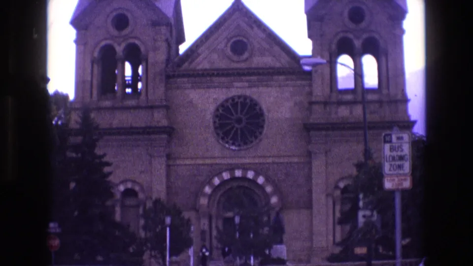 1973 New Mexico 8mm frame of a church front with twin bell towers, central rose window, striped arch detail over the entrance, trees, hills, and a bus loading zone sign.