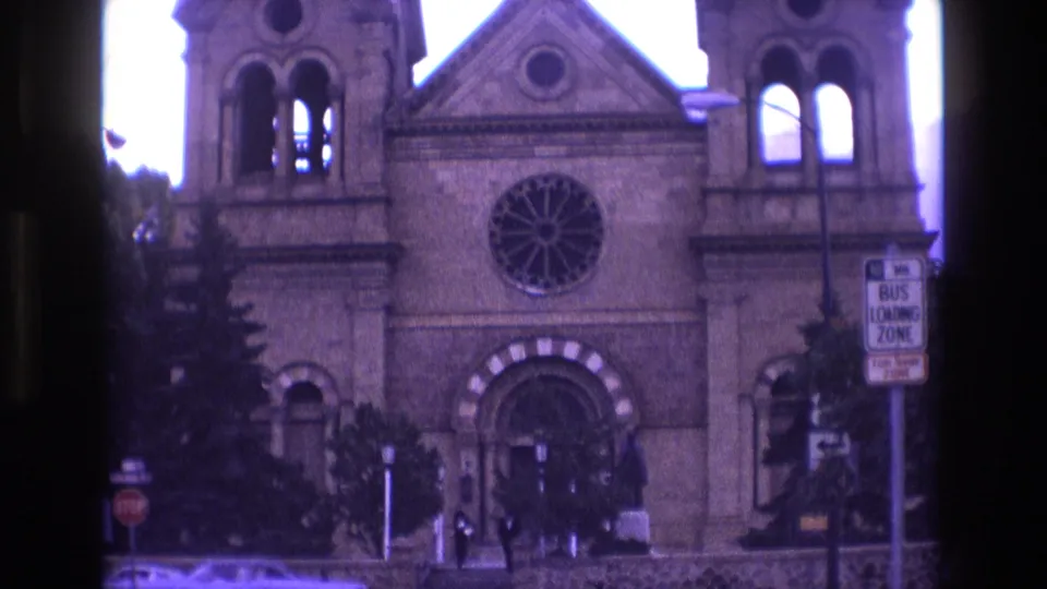 1973 New Mexico 8mm frame showing a large stone church exterior with twin bell towers, a rose window, pedestrians near the steps, street signage, and a car passing in front.