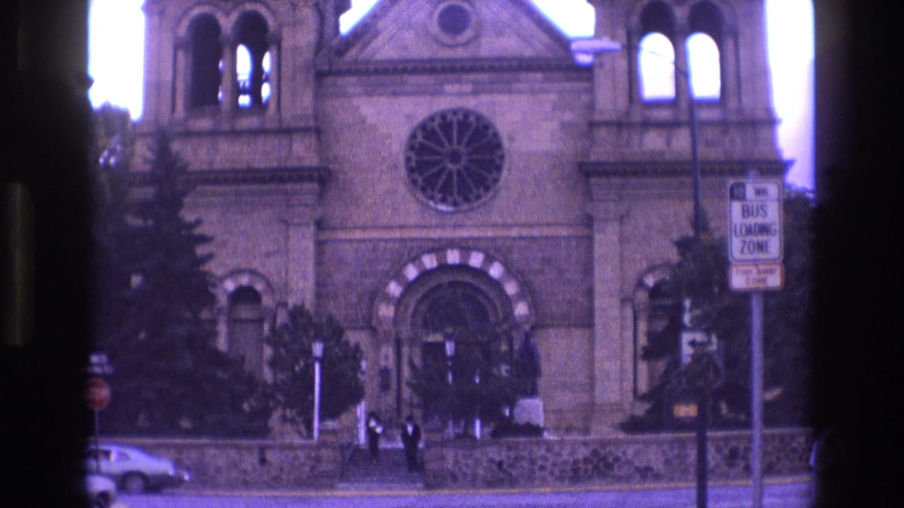 1973 New Mexico 8mm frame of the church entrance and rose window, with small pedestrians near the steps, a bus loading zone sign, trees, and a car on the street.