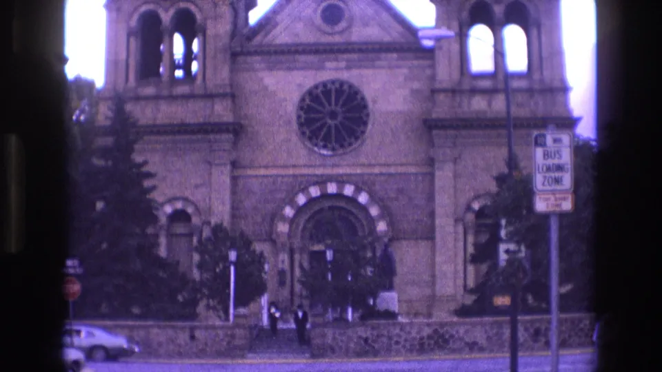 1973 New Mexico 8mm frame of the church entrance and rose window, with small pedestrians near the steps, a bus loading zone sign, trees, and a car on the street.