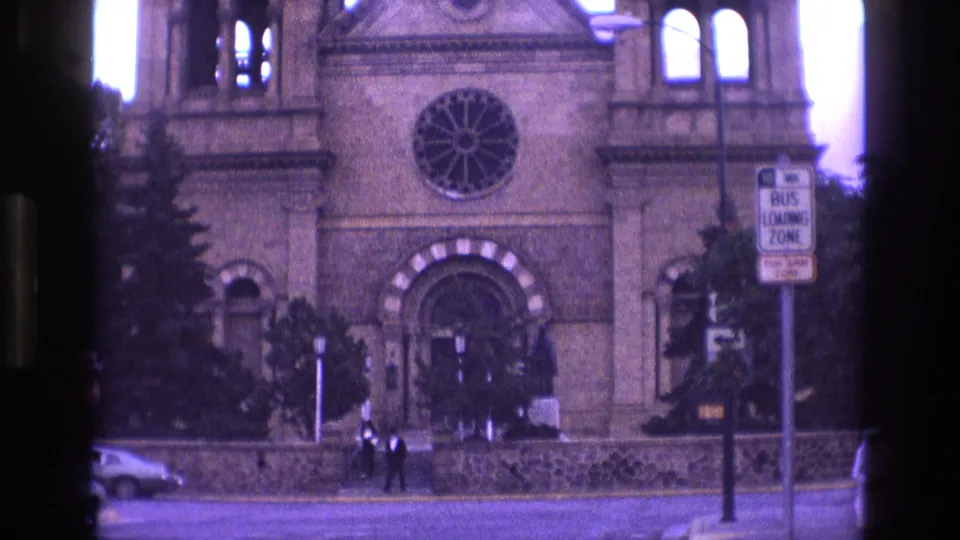 1973 New Mexico 8mm frame centered on the stone church facade, showing the rose window, arched entry, pedestrians by the steps, street signage, and a car partly visible at the curb.
