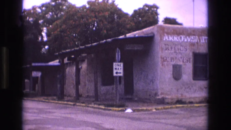 1973 New Mexico 8mm frame showing a nearby street corner with a one-way sign, low building, trees, curb, and partially readable wall lettering for an archdiocese-related office or site.