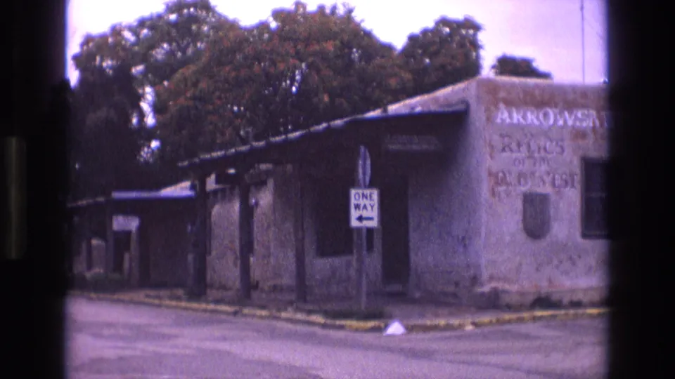 1973 New Mexico 8mm frame showing a quiet paved corner, a white one-way sign, trees, and faded lettering on a weathered adobe-style building.
